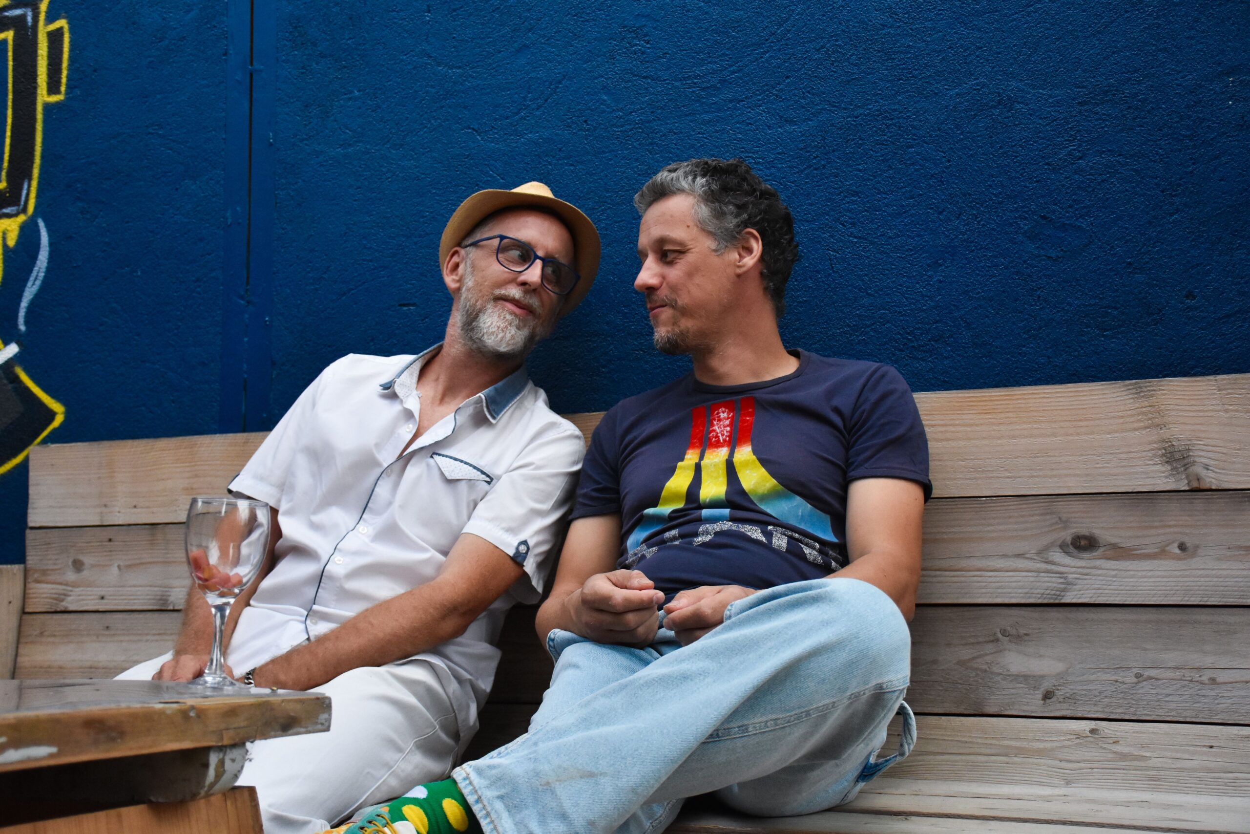 Deux hommes souriants, assis contre un mur bleu.